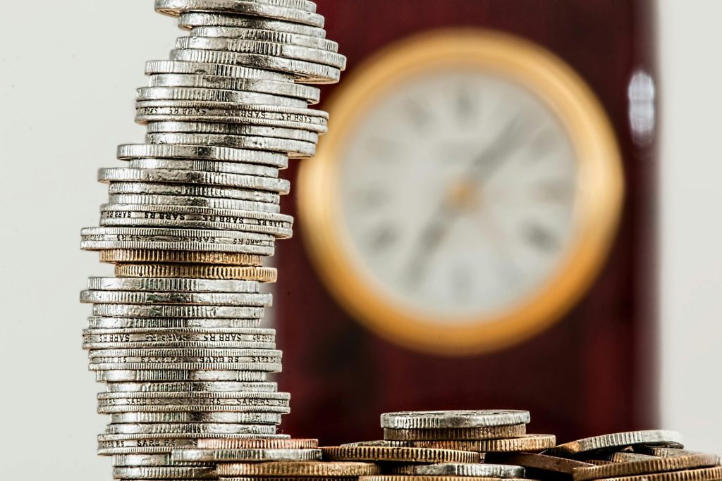 Eine Nahaufnahme gestapelter Münzen mit einer unscharfen Uhr, die die Beziehung zwischen Zeit und Geld symbolisiert.